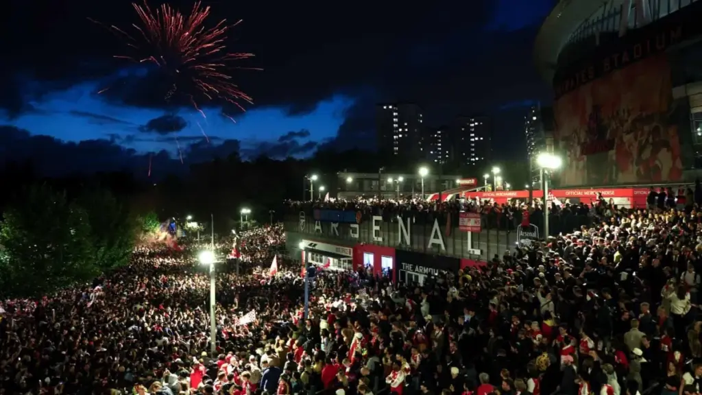 Arsenal Crowned Premier League Champions for First Time in 22 Years – The Wait is Over! 3 Arsenal players celebrating wildly with the Premier League trophy at the Emirates Stadium at night huge crowd in red and white Arsenal Crowned Premier League Champions for First Time in 22 Years – The Wait is Over!